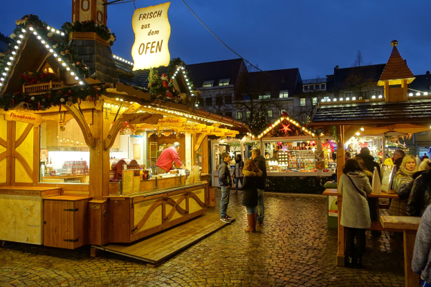 Ein belebter Weihnachtsmarkt auf einer Kopfsteinpflasterstraße bei Nacht mit Menschen um geschmückte Stände, Gebäuden im Hintergrund, Bäumen und einem klaren Himmel.