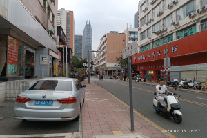 Ein Mann fährt mit einem Roller eine Straße in einer Stadt mit hohen Gebäuden entlang, mit Menschen auf den Bürgersteigen und Fahrzeugen auf der Straße, unter einem klaren blauen Himmel mit Bäumen im Hintergrund.