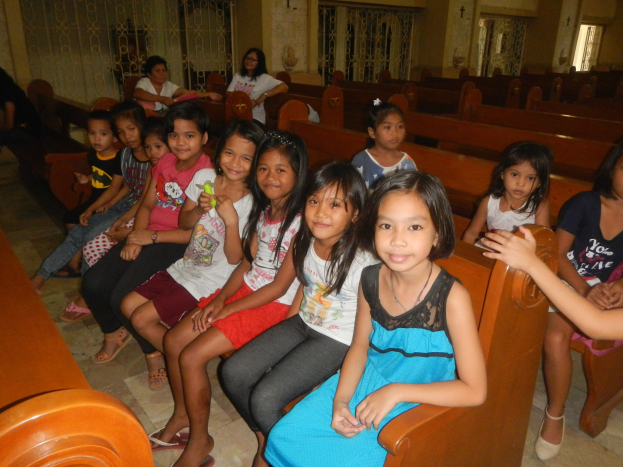Eine Gruppe junger Mädchen sitzt auf Kirchenbänken, mit ein paar Menschen im Hintergrund, umgeben von sichtbaren Wänden, Gittern und Fenstern.