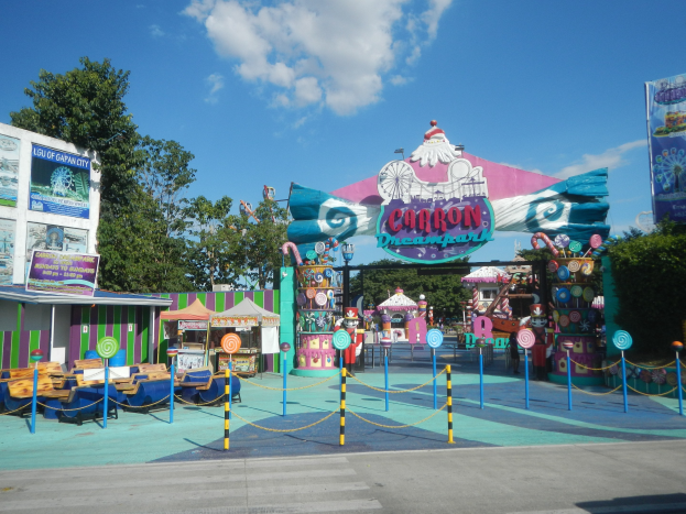 Karussell am Parkeingang mit Straße, Umzäunung, Ständen, Bannern, Bäumen und bewölktem Himmel im Hintergrund.