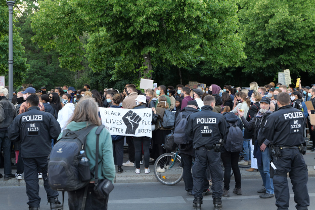 Eine große Gruppe von Menschen steht auf der Straße während einer Demonstration, einige halten Schilder und tragen Kappen und Taschen, mit einem Fahrrad im Vordergrund und Bäumen und einem Pfahl im Hintergrund.