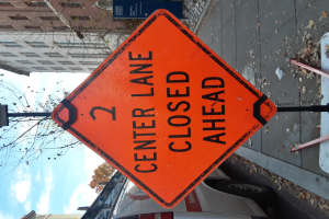 Ein oranges Schild am Straßenrand zeigt "2 Center Lane Closed Ahead", mit Fahrzeugen, Masten, Planken, Bäumen, Gebäuden und einem bewölkten Himmel im Hintergrund.