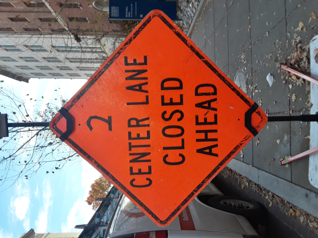 Ein oranges Schild am Straßenrand zeigt "2 Center Lane Closed Ahead", mit Fahrzeugen, Masten, Planken, Bäumen, Gebäuden und einem bewölkten Himmel im Hintergrund.