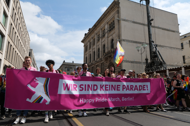 Eine Gruppe von Menschen, die auf der Straße in Berlin, Deutschland, mit einer rosa Schilder gehend, auf dem "Happy Pride March" steht, mit Gebäuden auf beiden Seiten der Straße und einem Fahnenmast im Vordergrund unter einem bewölktem Himmel.