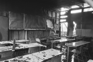 Schwarz-weißes Foto eines Klassenzimmers mit verkohlten Wänden, zerbrochenen Fenstern und verstreuten Tischen und Stühlen.