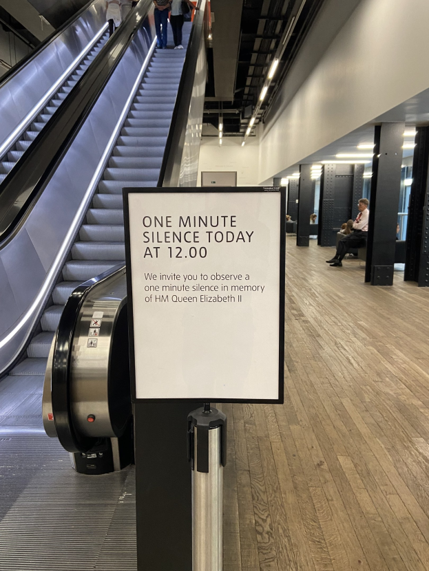 Eine Rolltreppe in einem Flughafen mit einem Schild, auf dem "Eine Minute Stille heute" steht, ein paar Menschen darauf und beleuchtete Decke im Hintergrund.