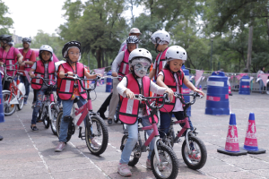Kinder mit Helmen fahren auf einer von Verkehrskegeln gesäumten Straße Fahrrad, einige mit Gesichtsbemalung, mit Bäumen und einem klaren blauen Himmel im Hintergrund.