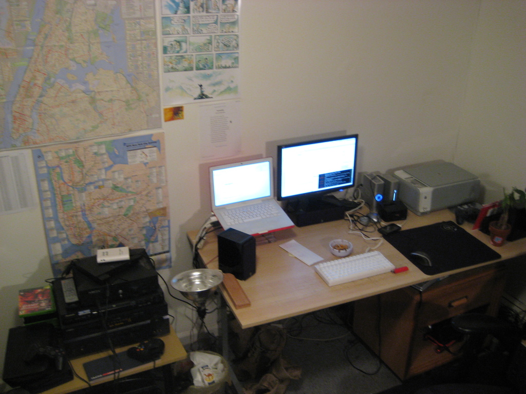 Ein Raum mit einem Tisch, auf dem ein Laptop, Lautsprecher, Monitor, Tastatur, Ball, Maus, Scanner und Papiere liegen; ein weiterer Tisch mit Elektronikgeräten im Vordergrund und eine Wand mit Charts und Postern im Hintergrund.