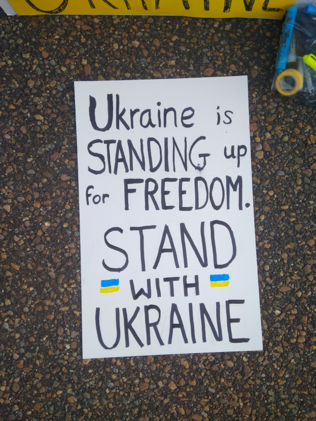 Weißer Papierbogen mit dem Text "Ukraine steht für Freiheit ein, steht mit Ukraine" auf dem Boden vor einer gelben Fahne, mit einer Plastikhülle rechts daneben, die verschiedene Gegenstände enthält.