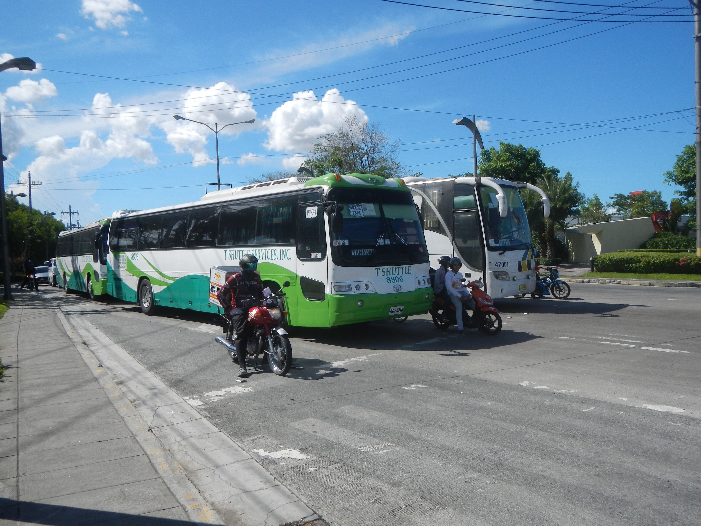 Ein grüner und weißer Shuttlebus steht am Straßenrand geparkt, vor ihm fahren Motorradfahrer, links ein grasbewachsener Fußweg und im Hintergrund Gebäude, Bäume, Laternenmasten und ein klarer blauer Himmel.