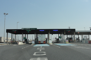 Eine Mautstelle mit einem vor ihr geparkten Lastwagen, neben einer Straße mit weißen und blauen Markierungen, Laternenmasten, Schildern und anderen Gegenständen, mit Gebäuden, Bäumen und einem klaren blauen Himmel im Hintergrund.