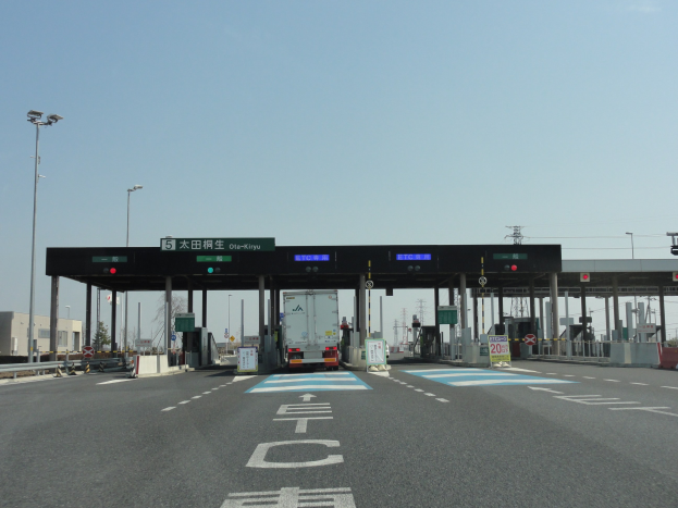 Eine Mautstelle mit einem vor ihr geparkten Lastwagen, neben einer Straße mit weißen und blauen Markierungen, Laternenmasten, Schildern und anderen Gegenständen, mit Gebäuden, Bäumen und einem klaren blauen Himmel im Hintergrund.