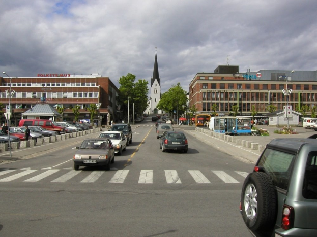 Stadtstraße mit parkenden Autos, Gebäuden mit Fenstern, Bäumen, Laternen und einem bewölkten Himmel im Hintergrund, mit einem sichtbaren Kfz-Kennzeichen.