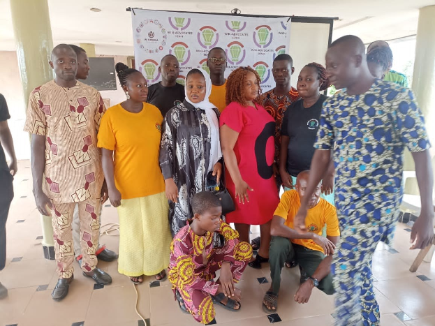 Eine Gruppe von Menschen vor einem Banner mit der Aufschrift "Advocates in Ghana: Ein Schritt-für-Schritt-Führer" mit Stühlen, Säulen, einem Fernseher und Deckenleuchten im Hintergrund.