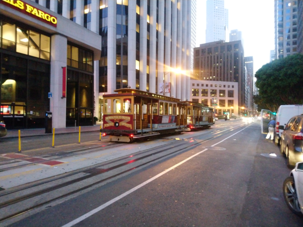 Seilbahn auf einer Stadtstraße in San Francisco mit Fahrzeugen, einem Fußgänger und einem geparkten Van, vor Buildings, Bäumen und Himmel.