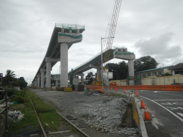 Baustelle mit einer Brücke im Hintergrund, eine Straße mit Absperrbaken auf der rechten Seite, Steine und Gras auf dem Boden, eine Bahnschiene auf der linken Seite, Bäume und Gebäude auf beiden Seiten der Straße und ein bewölkter Himmel oben.