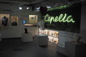 Ein Raum voller verschiedener Gegenstände, darunter Schaufensterpuppen mit Kleidern, Gegenstände auf Regalen, ein Display, eine Namensschilder, dekorative Elemente an der Decke und Deckenleuchten bei der Cepella-Ausstellung in Mailand.