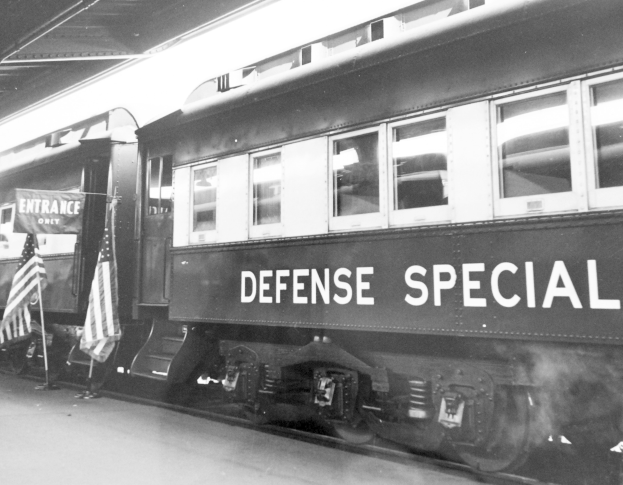 Schwarz-weißes Foto eines Zuges mit der Aufschrift "Defense Special" auf Gleisen unter einer Decke geparkt, mit zwei Flaggen links zu sehen.