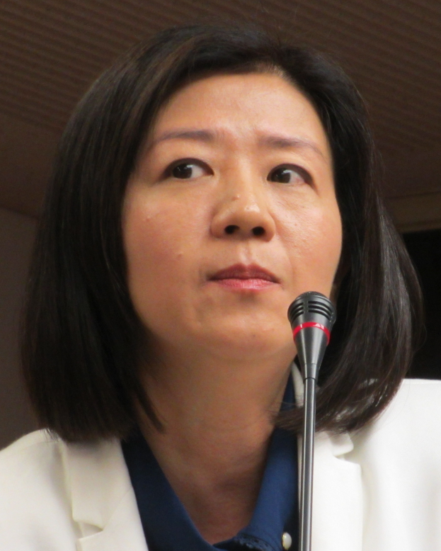 Eine Frau in einer weißen Jacke und Anzug spricht in ein Mikrofon vor einer Wand, mit einem ernsten Gesichtsausdruck.