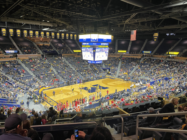 Eine große Arena mit Zuschauern, die ein Basketballspiel verfolgen, wobei einige stehen und andere sitzen, und eine Person mit einem Mobiltelefon in der Hand; die Decke hat Stangen und Lampen, und in der Mitte sind Tafeln mit dem Text "Michigan Wolverines Basketball" und ein Bildschirm.