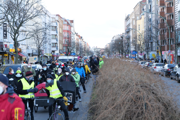 Eine große Gruppe von Menschen in Masken und Sicherheitswesten, die auf Fahrrädern eine von Bäumen gesäumte Straße entlangfahren, mit Gebäuden, Fahrzeugen, Laternenmasten und Texttafeln, unter einem klaren blauen Himmel.