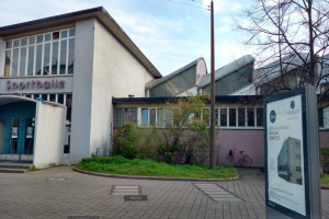 Ein großes Gebäude mit einem Schild davor, umgeben von Bäumen, Pflanzen, Gras, Fahrrädern und einem Pfahl, mit einem bewölkten Himmel im Hintergrund, identifiziert als das Sportballe, das 2020 eröffnet werden soll.