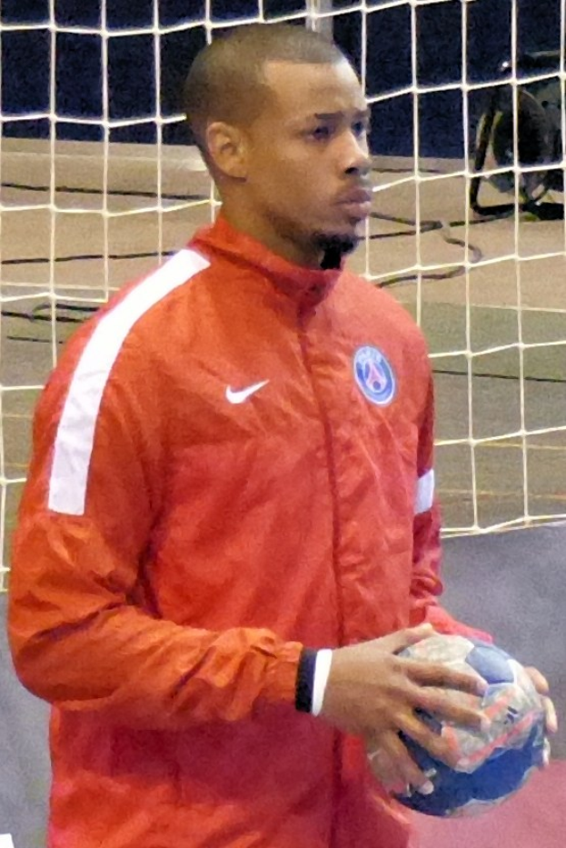 Ein Mann in einem blauen T-Shirt und Shorts steht vor einem Tor und hält einen Fußball, mit entschlossener Miene und konzentriertem Blick.