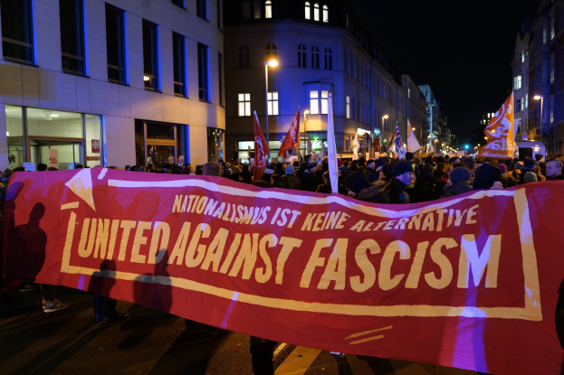Eine Gruppe von Menschen marschiert mit einem "Einheit gegen Faschismus"-Schild die Straße entlang, mit Gebäuden, Laternen und Schildern an der Straße und einem sichtbaren Himmel im Hintergrund.