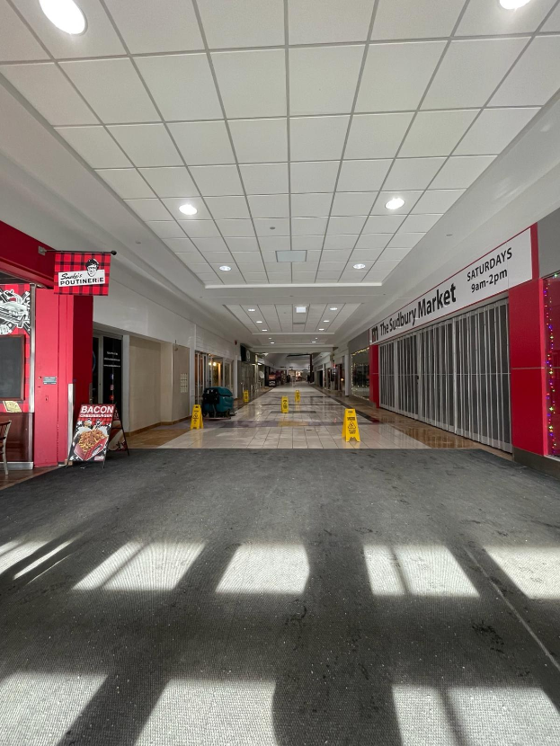 Innenansicht eines Einkaufszentrums mit Deckenbeleuchtung, einem Stuhl und einer Tafel, auf der "Sydbury Market - Vorher und Nachher" steht, auf der linken Seite und verstreuten Gegenständen auf der rechten Seite und dem Boden.