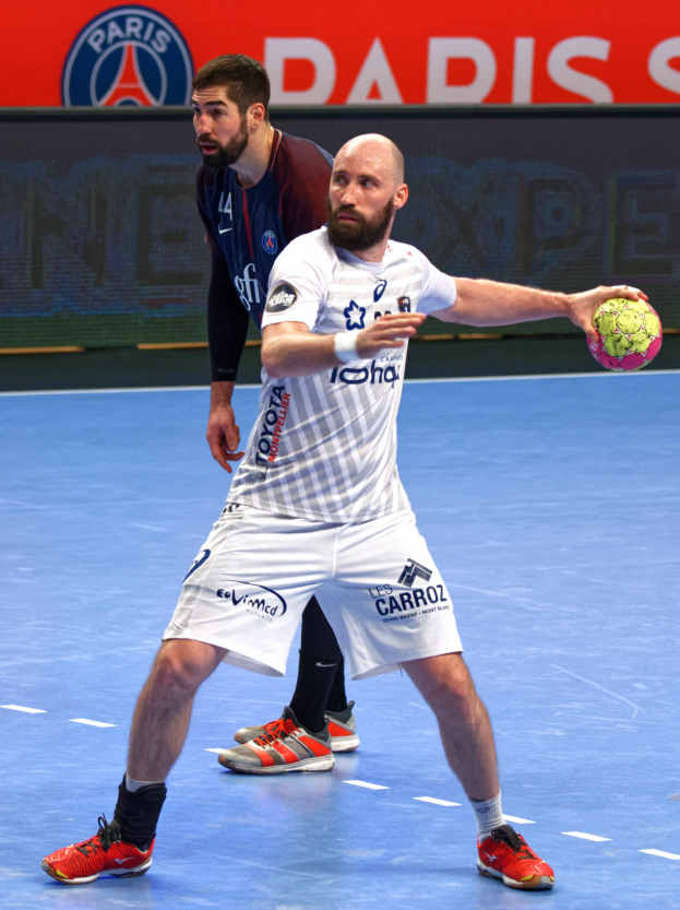 Zwei Männer beim Handballspielen auf einem Platz, einer hält den Ball, im Hintergrund eine Tafel mit 'Paris Saint-Germain vs Paris Saint-Germain'.