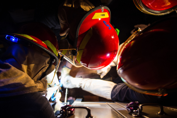 Feuerwehrleute in Helmen und Handschuhen arbeiten an einem Feuerwehrauto in einem schwach beleuchteten Bereich, mit einem Tisch mit Vorräten auf der rechten Seite.