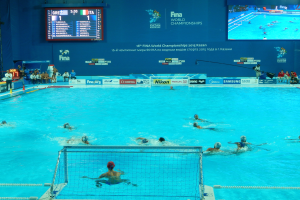Eine Gruppe von Menschen, die Wasserball in einem großen Pool mit einem Netz in der Mitte spielen, Zuschauer auf der linken Seite und ein Bildschirm mit Text und Logos im Hintergrund bei den 2016 FINA World Championships in Kazan.