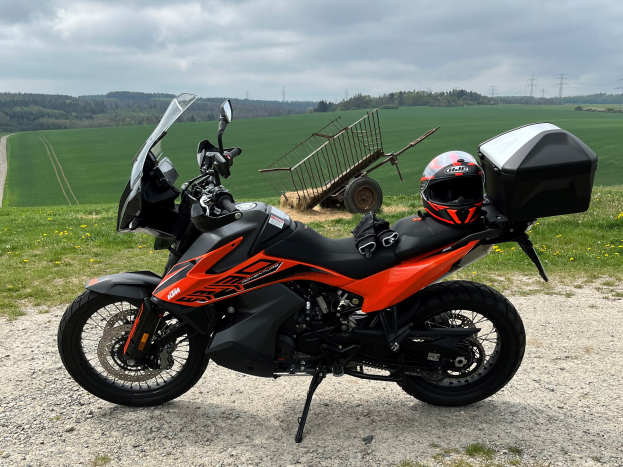 Eine KTM 1290 Super Adventure R Motorrad auf einem Schotterweg geparkt mit einem Helm und einem Gepäckträger daneben, umgeben von grünem Gras, hohen Bäumen und Strommästen unter einem bewölkten Himmel.