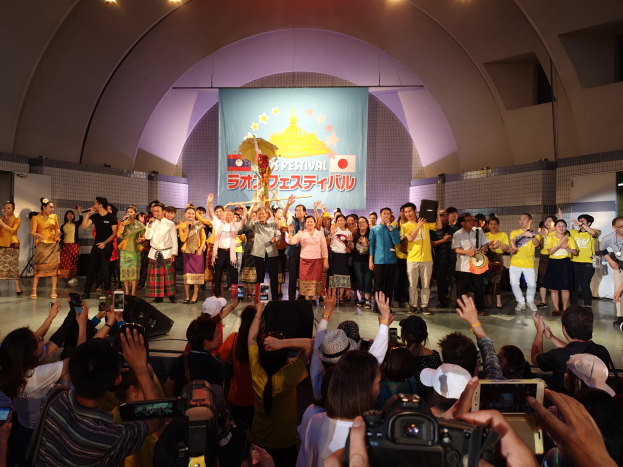 Gruppe von Menschen auf einer Bühne mit Kameras, Handys und Musikinstrumenten während einer Festival-Eröffnung mit Bannern, Text und Deckenbeleuchtung im Hintergrund.