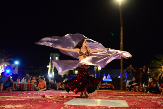 Eine Frau in einem Bauchtanzkostüm tanzt auf einem roten Teppich, während eine Menge mit Kameras zusieht, mit Bäumen, Lichtern, einem Pfahl und Himmel im Hintergrund.