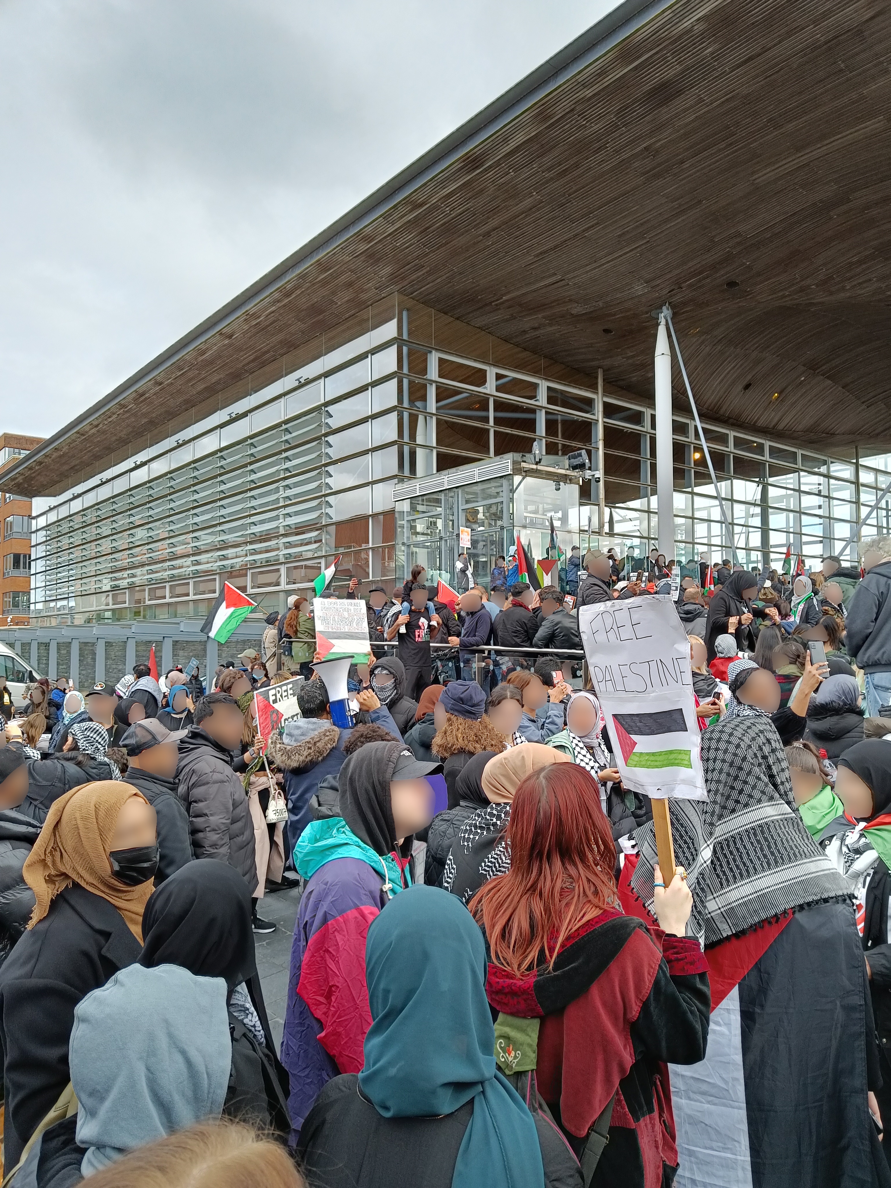 Eine große Gruppe von Menschen steht vor einem Gebäude und hält Schilder und Fahnen hoch, während ein Fahrzeug auf der linken Seite und der Himmel oben zu sehen ist.