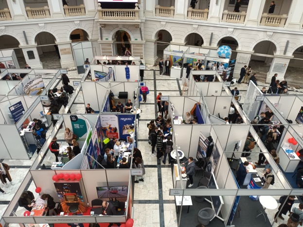 Eine große Gruppe von Menschen versammelt sich in einem Raum mit Ständen, Bannern, Tischen, Stühlen, Ballons und einem Gebäude mit Bögen, Säulen, Geländern und einem Bildschirm im Hintergrund.
