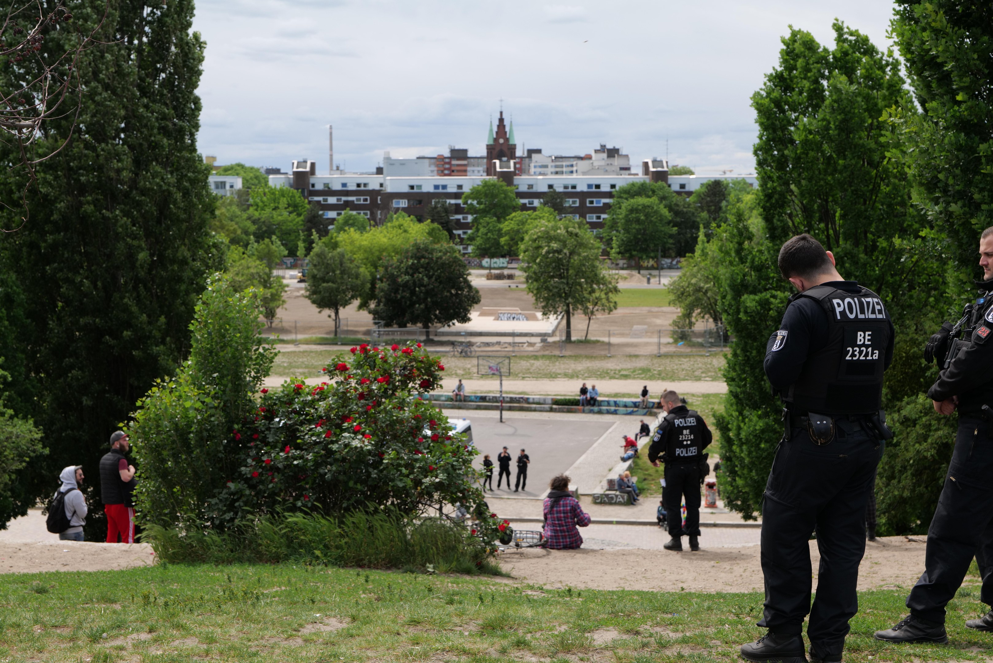 Zwei Polizeibeamte vor einer Gruppe von Menschen in einem Park mit saftigem Grün, Bäumen, bunten Blumen, Gebäuden, Pfählen und einem klaren blauen Himmel.