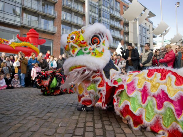 Eine farbenfrohe chinesische Neujahrsfeier in Amsterdam mit einem Löwen tanzen im Vordergrund und einer Menschenmenge, einige halten Kameras, vor einem Hintergrund aus Gebäuden, Laternenmasten und einem klaren blauen Himmel.