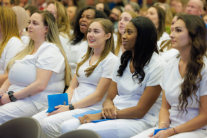 Eine Gruppe von Frauen in weißen Kleidern sitzt in einem Hörsaal, jede hält ein blaues Objekt, mit einem unscharfen Hintergrund, der die Frauen betont, die scheinbar Teil eines Pflegeprogramms sind.