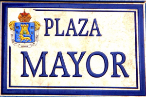 Rechtwinkliges weißes Schild mit schwarzer Schrift "Plaza Mayor" in fetter Schrift, blauem Rand und einem gekrönten Wappen-Logo an der Seite eines Gebäudes.