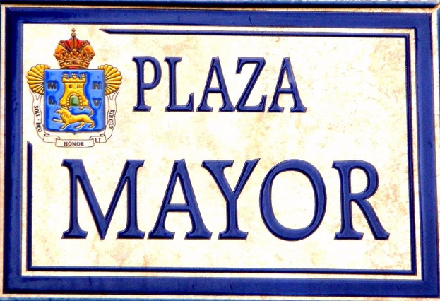 Rechtwinkliges weißes Schild mit schwarzer Schrift "Plaza Mayor" in fetter Schrift, blauem Rand und einem gekrönten Wappen-Logo an der Seite eines Gebäudes.