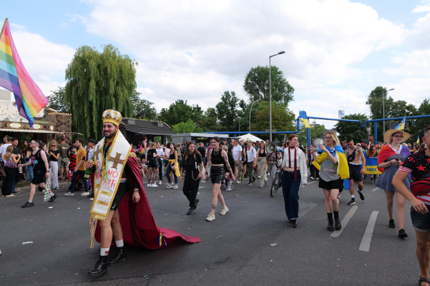 Eine Gruppe von Menschen marschiert bei der Gay Pride Parade 2018 mit einer Regenbogenfahne und Musikinstrumenten, im Hintergrund sind Laternenpfähle, Bäume und Schuppen zu sehen.