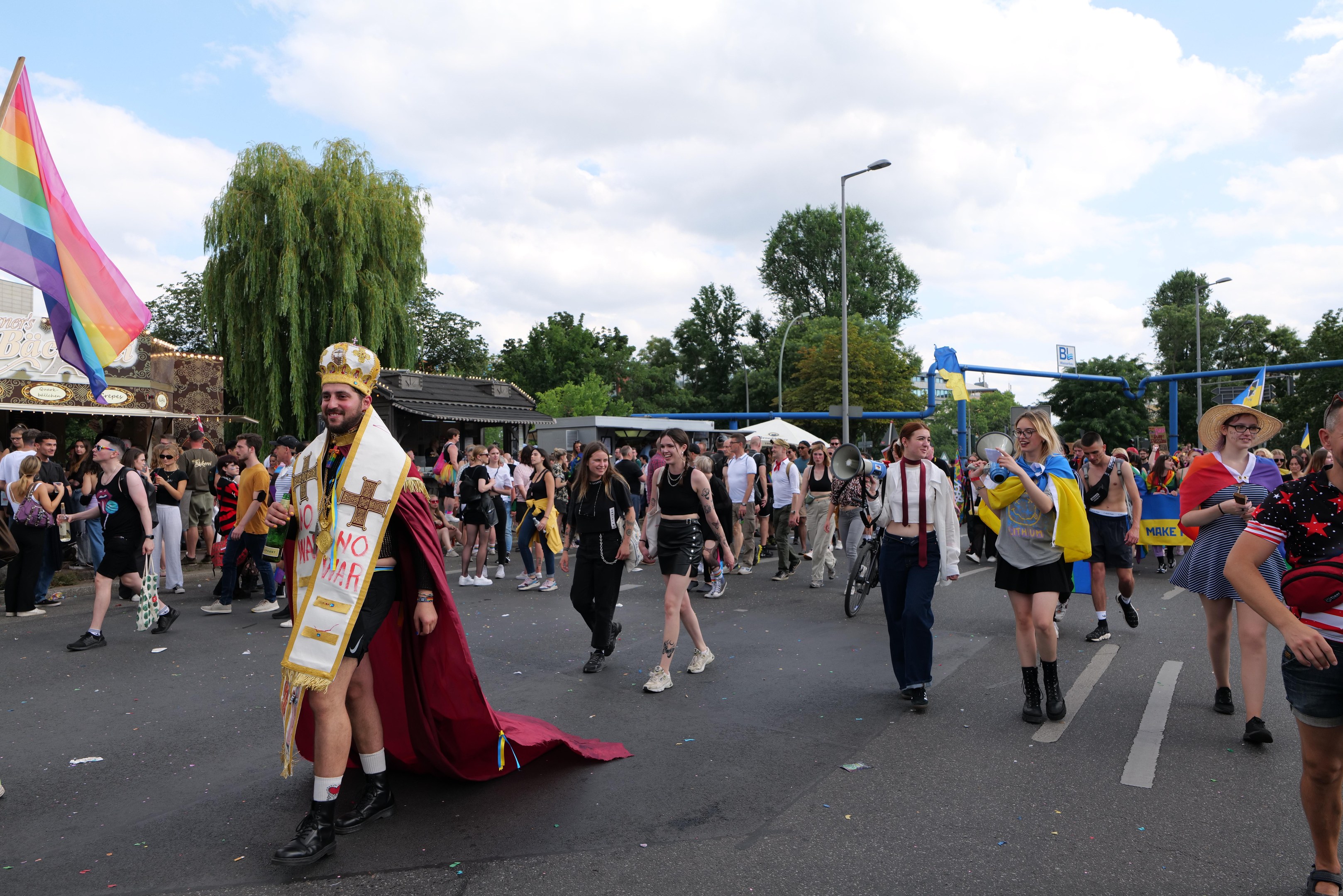 Eine Gruppe von Menschen marschiert in einem Umzug, einige halten Musikinstrumente und andere tragen Mützen, tragen eine Regenbogenflagge, mit Laternenmasten, Bäumen, Hütten und einem bewölkten Himmel im Hintergrund.