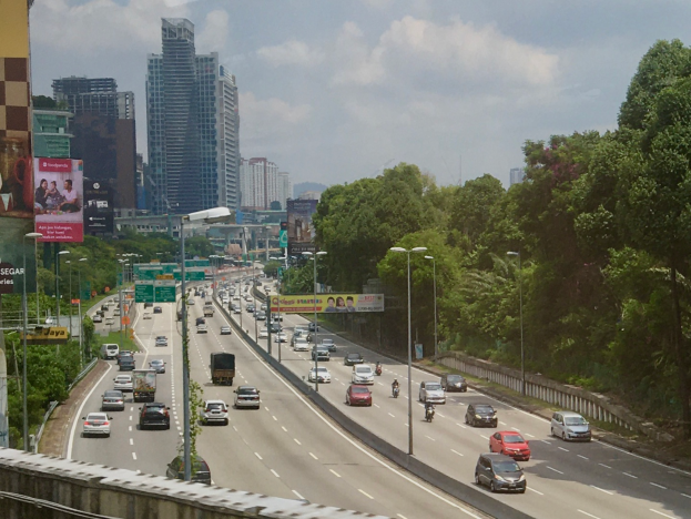 Eine vielbefahrene Autobahn mit zahlreichen Fahrzeugen, gesäumt von hohen Gebäuden, Straßenlaternen, Werbetafeln und Bäumen unter einem bewölkten Himmel.