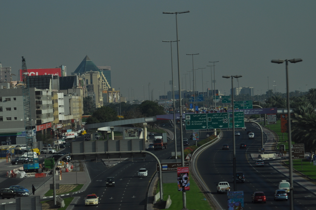 Luftaufnahme einer Stadt mit Gebäuden, Türmen, Kraftfahrzeugen, Werbetafeln, Schildern, Straßen, Bäumen, Straßenlaternen, Menschen, Verkehrskegeln und Informationsanzeigen unter einem klaren Himmel.