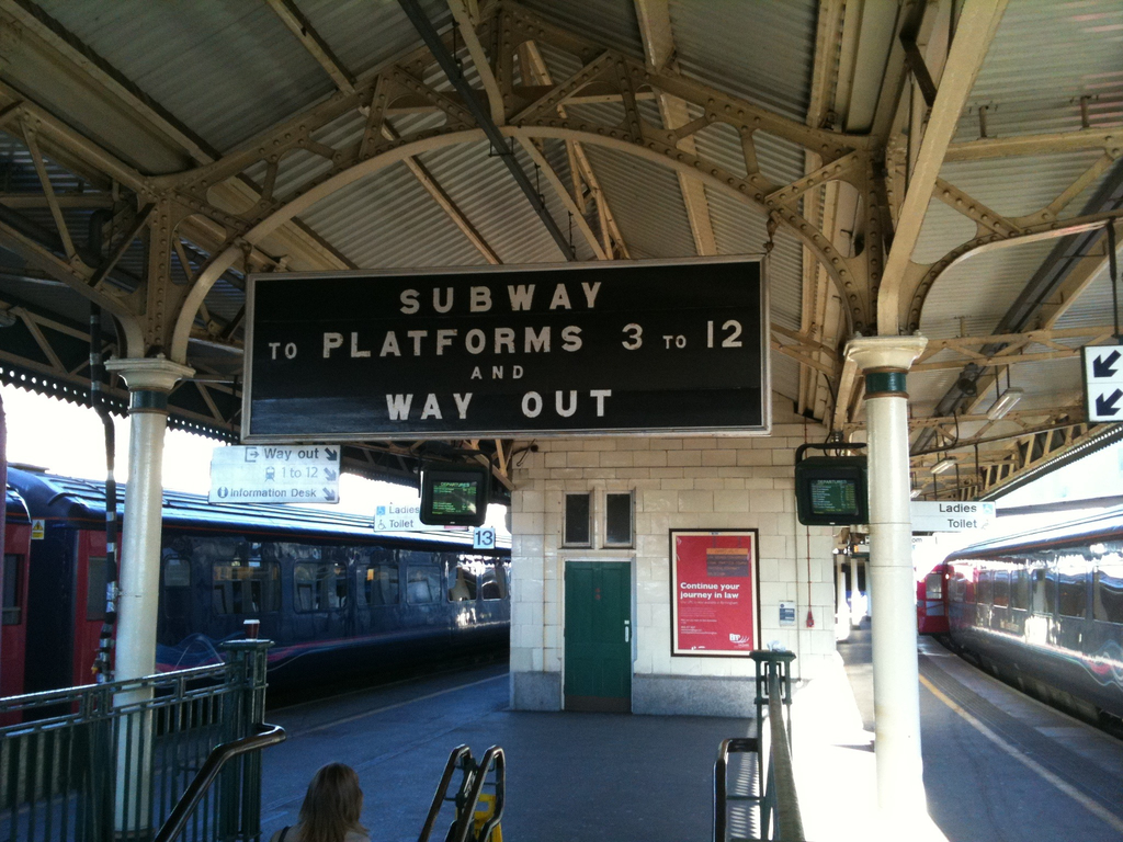Zwei Züge stehen neben einem Bahnsteig mit zwei Bildschirmen und mehreren Schildern, die von der Decke hängen, eine Person ist in der Nähe einer Treppe und eine Tür ist an der Wand sichtbar.