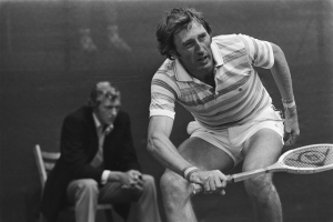 Mann schwingt Tennisracket gegen einen Ball, im Hintergrund sitzt jemand auf einem Stuhl, dargestellt in Schwarz-Wei├č.