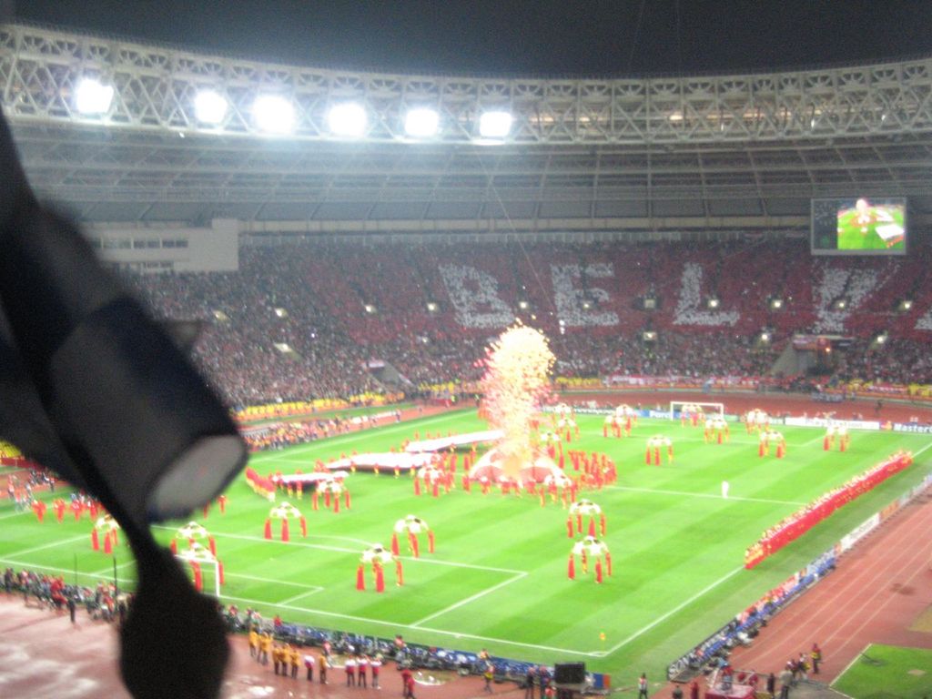 Ein Stadion mit Flutlicht, Eisengittern, Zuschauern, stehenden Personen, Werbetafeln, Bildschirmen, Überwachungskameras, Flaggen und Dekorationen.