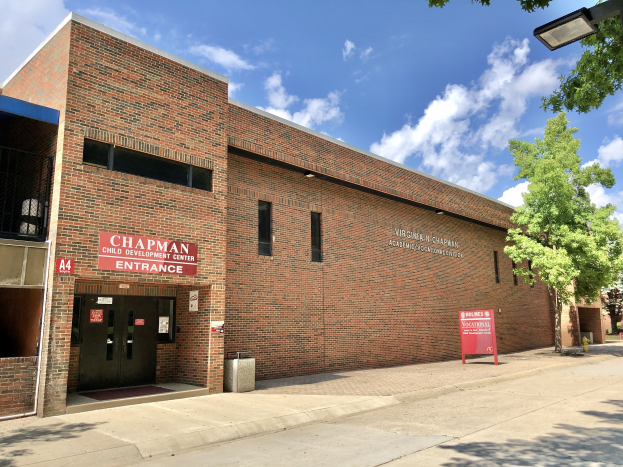 Außenansicht eines Backsteingebäudes namens 'Chapman Child Development Center' mit Fenstern, Türen, einer Namenstafel, einem Mülleimer, einem Straßenpfahl, Bäumen, parkenden Fahrzeugen und einem bewölkten Himmel.